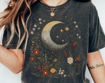 May include: Dark gray t-shirt featuring a celestial design. The graphic includes a crescent moon, stars, and various wildflowers in shades of orange, yellow, and white. The shirt has a vintage, worn look.