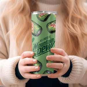 Puede incluir: Un vaso verde con un patr&oacute;n repetido de latas de Mountain Dew y el texto "All I need is Mountain Dew". El vaso tiene una tapa plateada y lo sostiene una persona.