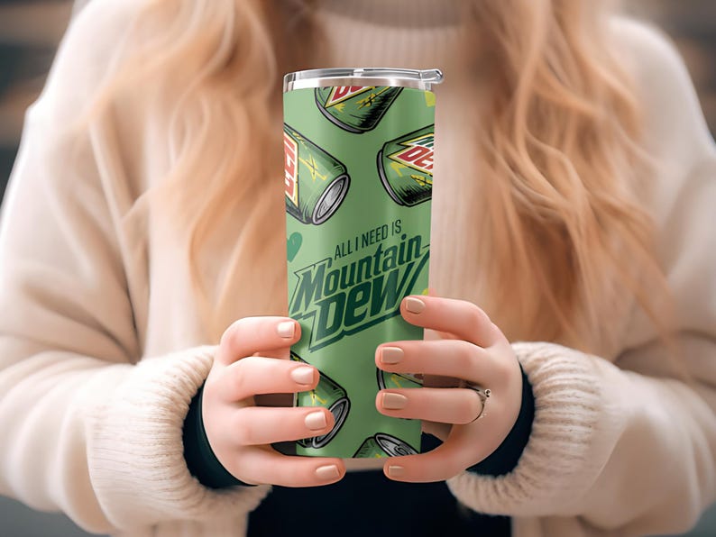 Puede incluir: Un vaso verde con un patr&oacute;n repetido de latas de Mountain Dew y el texto "All I need is Mountain Dew". El vaso tiene una tapa plateada y lo sostiene una persona.