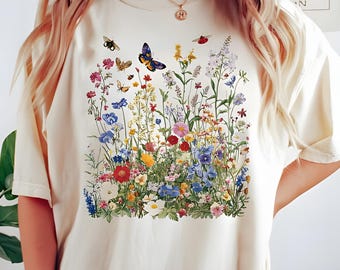 May include: Cream-colored t-shirt featuring a vibrant floral design with butterflies, bees, and ladybugs. The graphic showcases a colorful array of wildflowers in shades of red, blue, yellow, and white, creating a cheerful, nature-inspired aesthetic.