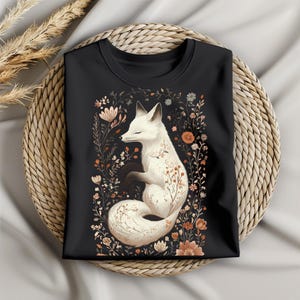 May include: Black crew neck t-shirt featuring a white fox illustration surrounded by floral elements in shades of orange, brown, and beige. The fox is curled up with its eyes closed. The shirt is folded on a woven basket.