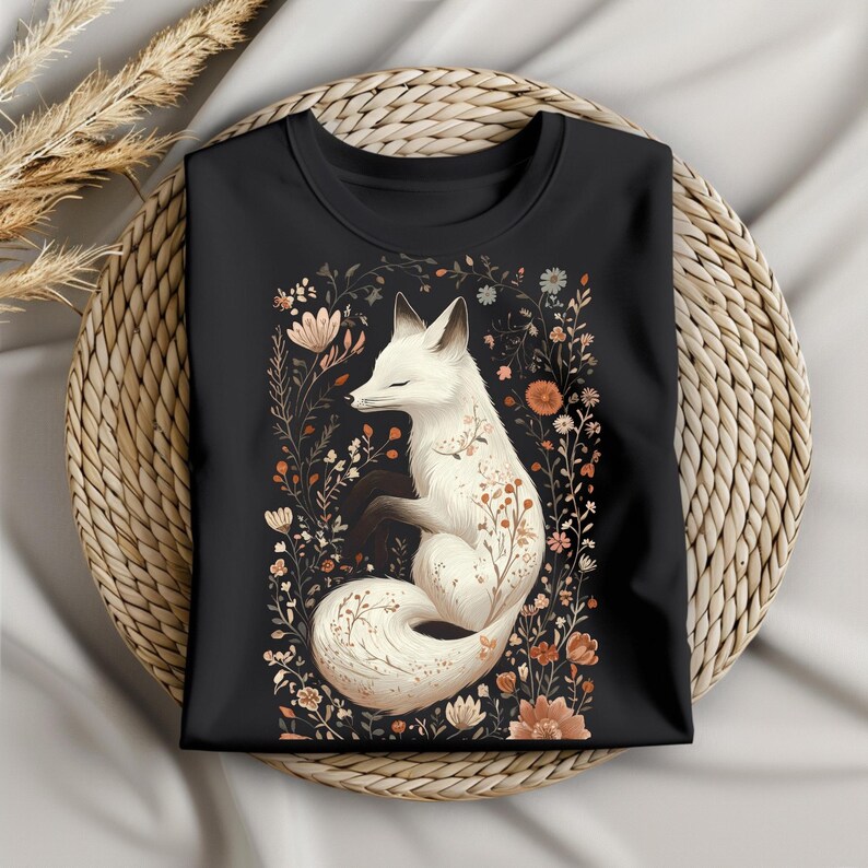 May include: Black crew neck t-shirt featuring a white fox illustration surrounded by floral elements in shades of orange, brown, and beige. The fox is curled up with its eyes closed. The shirt is folded on a woven basket.