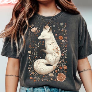 May include: Dark gray t-shirt featuring a white fox illustration surrounded by flowers. The fox has closed eyes and is adorned with floral details. The design includes various flowers in shades of brown, orange, and white.