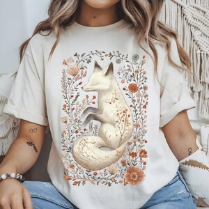 May include: Cream-colored t-shirt featuring a serene white fox illustration surrounded by a floral wreath. The fox has closed eyes and is nestled among flowers in shades of orange, brown, and cream. The design is centered on the shirt.