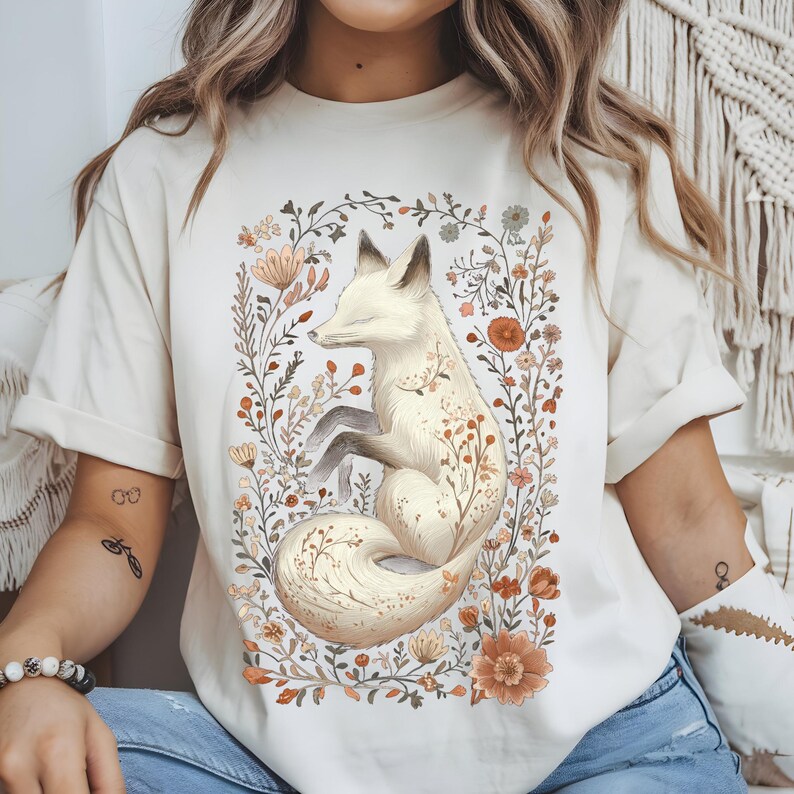 May include: Cream-colored t-shirt featuring a serene white fox illustration surrounded by a floral wreath. The fox has closed eyes and is nestled among flowers in shades of orange, brown, and cream. The design is centered on the shirt.