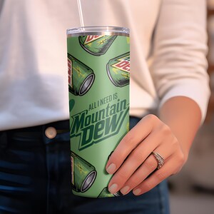 Puede incluir: Un vaso verde con un patr&oacute;n repetido de latas de Mountain Dew y el texto "ALL I NEED IS Mountain Dew". El vaso tiene una pajita blanca y una tapa plateada. El dise&ntilde;o est&aacute; sobre un fondo verde claro.