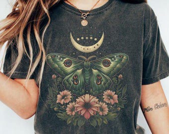 May include: A dark gray t-shirt with a green moth and floral design. The moth has a crescent moon on its back and the design includes a crescent moon with stars.