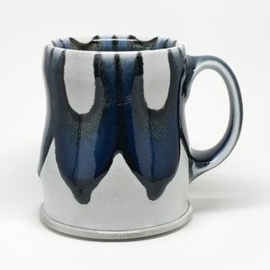 Op de afbeelding: Een witte keramische mok met een blauwe en zwarte glazuur. De glazuur heeft een druppel effect, waardoor een uniek patroon ontstaat.
