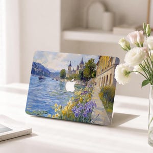 May include: A laptop skin featuring an impressionist-style painting of a lakeside scene with blue water, yellow and purple flowers in the foreground, a stone pathway, yellow buildings, and a distant church under a cloudy sky with an Apple logo cutout.