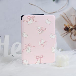 Può includere: Una custodia rosa per e-reader con un motivo di coniglietti bianchi e fiocchi.
