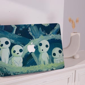 Può includere: Una custodia per laptop con una scena di foresta con quattro figure bianche, simili a fantasmi, con occhi neri. Lo sfondo è una foresta blu scuro e verde con un logo Apple bianco. Le figure sono su un'area erbosa verde.