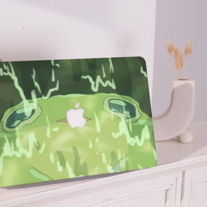 May include: A green laptop cover with a cartoon frog design and an Apple logo. The frog has large eyes and is set against a green background with white accents. The cover is on a white surface.