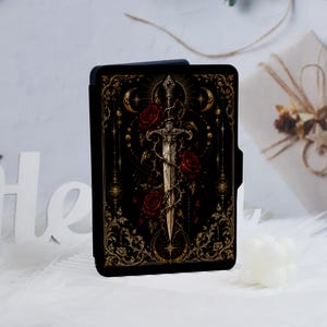 May include: A black e-reader case featuring a central silver sword entwined with thorny vines and deep red roses. The design includes gold celestial motifs, including crescent moons and sunbursts, set against a dark background with ornate filigree.