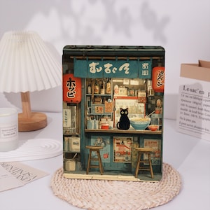 May include: A rectangular metal sign featuring a detailed illustration of a Japanese izakaya shop front. A black cat sits on the counter next to a bowl of noodles. The shop has blue signage with white Japanese text, red lanterns, and two wooden stools.
