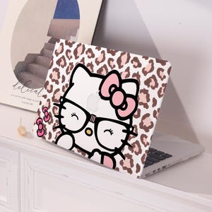 May include: A white laptop case featuring a Hello Kitty design with glasses and a pink bow. The case has a leopard print background in shades of brown and tan. The laptop is open on a white surface.