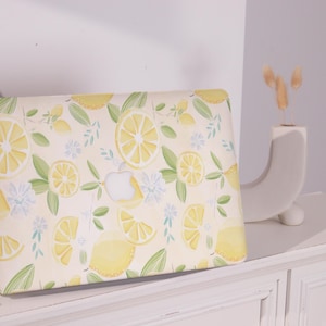 Può includere: Una custodia per laptop con un motivo a limoni. La custodia presenta un design ripetuto di limoni gialli, foglie verdi e fiori bianchi su uno sfondo color crema. Il logo Apple è visibile.