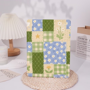 May include: An iPad case with a patchwork design featuring a variety of colours and patterns, including green, blue, yellow, and white. The case has a floral pattern, a checkered pattern, and a star.