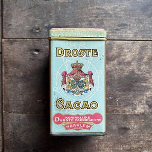 Könnte beinhalten: Eine Vintage-Blechdose für Droste Cacao, eine niederländische Marke für Kakaopulver. Die Dose ist blau mit goldener Verzierung und zeigt ein königliches Wappen mit einer Krone und zwei Löwen. Der Text auf der Dose lautet "Droste Cacao, Koninklijke Droste Fabrieken NV, Haarlem Holland."