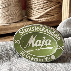 Könnte beinhalten: Eine ovale, grüne Dose mit dem Text "Stahlstecknadeln Maja" in weißen Buchstaben. Die Dose trägt auch den Text "50 Gramm No.". Die Dose liegt auf einer grauen, strukturierten Oberfläche. Zwirnspulen sind im Hintergrund.