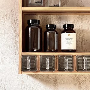 Könnte beinhalten: Ein Holzregal mit drei dunkel bernsteinfarbenen Glasflaschen mit schwarzen Deckeln, von denen eine ein weißes Etikett mit der Aufschrift OMEGA 3 COMPLETE DHA EPA ALA trägt. Darunter befinden sich vier klare Glasschubladen mit vertikaler Riffelung.