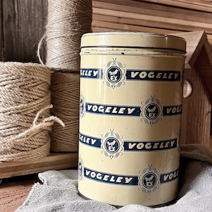 Könnte beinhalten: Eine cremefarbene, zylindrische Dose mit dem Wort "VOGELEY" in Marineblau, das sich um den Körper wiederholt. Die Dose hat ein Vogel-Emblem und befindet sich neben Garnrollen. Im Hintergrund befindet sich ein Vogelhaus aus Holz.