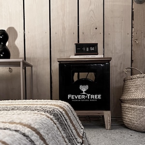Könnte beinhalten: Ein schwarzer Nachttisch aus Holz mit dem Logo "FEVER-TREE PREMIUM NATURAL MIXERS". Der Nachttisch hat eine Digitaluhr, die die Uhrzeit 8:54 anzeigt. Ein geflochtener Korb und ein Bett mit einer gestreiften Decke sind ebenfalls zu sehen.