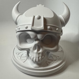 Puede incluir: Escultura de calavera de cerámica blanca con un casco vikingo con cuernos. La calavera está sentada sobre una base redonda con un diseño de nudo celta.