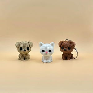 May include: Three small, knitted animal figurines: two dogs and a cat. The dogs are tan and brown, and the cat is white. The figurines have black eyes and pink noses. The brown dog has a keychain attached.