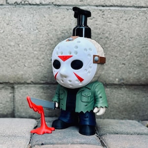 May include: A soap dispenser shaped like a character wearing a white hockey mask with red accents. The figure wears a green jacket and dark pants, holding a grey knife with a red liquid effect dripping from the blade onto the base surface.