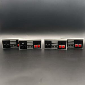 May include: Four rectangular, gray and black controller-shaped objects with red buttons and a directional pad. The objects are arranged horizontally on a dark surface, evoking a retro video game theme.