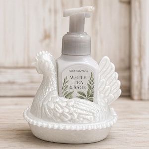 May include: A white ceramic chicken-shaped soap holder containing a square plastic bottle of Bath & Body Works White Tea & Sage foaming hand soap. The holder features a textured feather design and a beaded rim base with a pump dispenser.