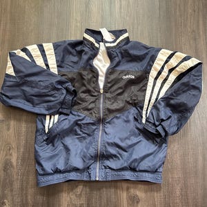 May include: Navy blue Adidas track jacket with white stripes on the sleeves. The jacket has a black panel across the chest and the Adidas logo on the chest. It features a full zip and ribbed cuffs and hem.
