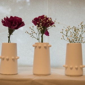 Könnte beinhalten: Drei weiße Keramikvasen mit einer Reihe kleiner Noppen am Boden. Die Vasen sind mit rosa Nelken und weißen Babyschnabel gefüllt.