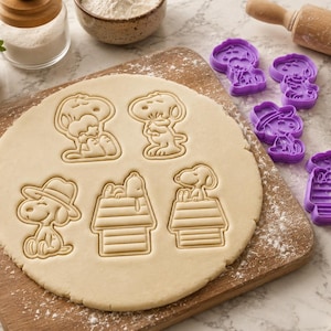 May include: A set of purple plastic cookie cutters featuring Snoopy in various poses, including sitting, wearing a hat, and resting on a doghouse. The cutters are shown alongside a rolled-out sheet of cookie dough with matching embossed designs.