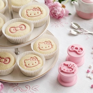 Puede incluir: Una bandeja de dos niveles exhibe cupcakes con diseños de lazo rosa y Hello Kitty. Un juego de té rosa, cucharas y flores complementan los dulces. También hay sellos rosas con diseños de lazo y Hello Kitty.