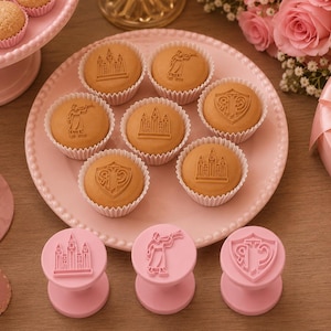 Könnte beinhalten: Eine Anordnung von Backwaren und Dekorationen in Rosatönen. Das Bild zeigt hellbraune Gebäckstücke mit geprägten Designs auf einem rosa Teller. Rosa Stempel mit passenden Designs sind ebenfalls vorhanden, zusammen mit einem rosa Kuchenständer und einer herzförmigen Geschenkbox.