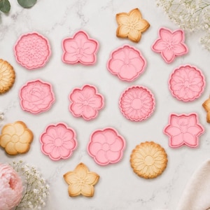 May include: A collection of pink plastic flower-shaped cookie cutters arranged alongside several baked, light brown cookies with intricate floral patterns pressed into their surfaces, featuring various petal designs and circular shapes.