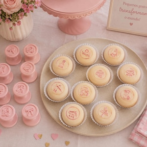 May include: A selection of pink stamps with different designs, and a plate of round pastries, each stamped with a corresponding design. A pink cake stand and a vase of pink roses are also visible. The text in the image says "Pequenos gestos transformam o mundo!"