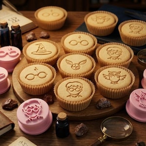 Peut inclure: Des biscuits décorés avec des motifs tels que des lunettes et un hibou, accompagnés de tampons roses assortis. De petites bouteilles en verre et une loupe sont également présentes, le tout disposé sur une surface en bois.