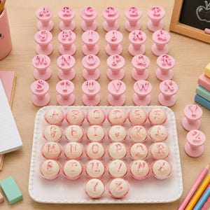 Könnte beinhalten: Rosa Alphabetstempel und Cupcakes mit Buchstaben auf einem weißen Tablett. Die Stempel sind in Reihen angeordnet, und die Cupcakes befinden sich in einem Raster. Im Hintergrund befindet sich eine kleine Tafel mit einer Apfelzeichnung.