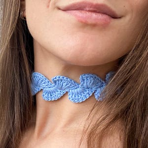 Può includere: Una collana girocollo all'uncinetto azzurra. La collana presenta un motivo a onde ripetuto. Il girocollo è indossato attorno al collo. Lo sfondo è di colore grigio chiaro.