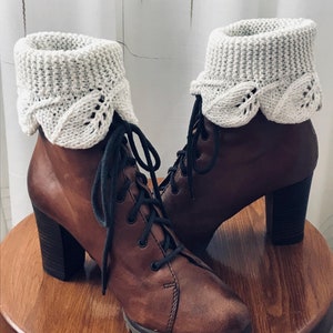 Könnte beinhalten: Braune Lederstiefel mit Knöchelhöhe, klobigem Absatz und weißen gestrickten Stiefelmanschetten. Die Manschetten haben einen gewellten Rand und ein zartes Blattmotiv.