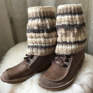 Könnte beinhalten: Braune Lederstiefel mit gestrickten Beinwärmern. Die Beinwärmer sind beige, braun und weiß mit einem strukturierten Muster. Die Stiefel haben Schnürsenkel und liegen auf einer weißen, flauschigen Oberfläche.