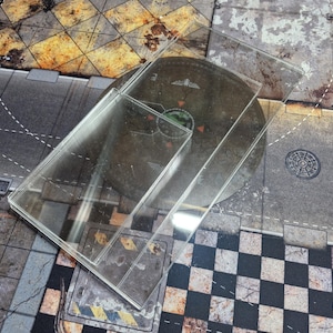 Może przedstawiać: Przezroczysta nakładka na planszę do gry z akrylu o prostokątnym kształcie, umieszczona na okrągłej planszy do gry. Nakładka ma chronić planszę do gry. Tło przedstawia szczegółową scenę uliczną z wzorem w szachownicę.