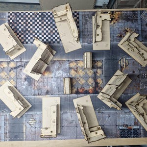 Può includere: Una collezione di elementi di terreno in legno tagliati al laser per giochi da tavolo, tra cui facciate di edifici e muri, disposti su un tappetino di gioco dettagliato con un design di paesaggio urbano. I pezzi sono incompiuti e pronti per la verniciatura.