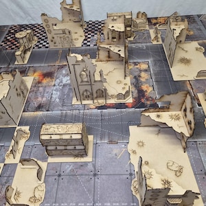 May include: A collection of laser-cut wooden miniature terrain pieces arranged on a printed gaming mat. The set includes various ruined building structures, wall sections, and a rectangular chest, all featuring intricate engraved details.