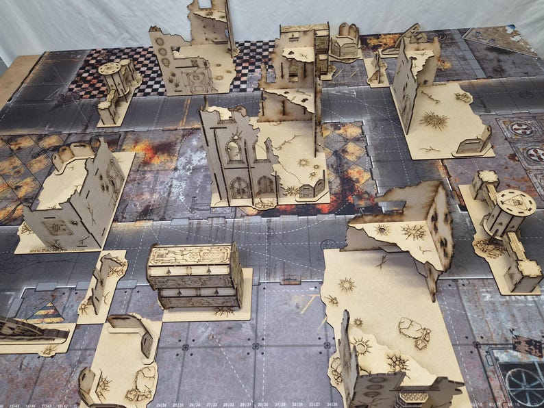 May include: A collection of laser-cut wooden miniature terrain pieces arranged on a printed gaming mat. The set includes various ruined building structures, wall sections, and a rectangular chest, all featuring intricate engraved details.
