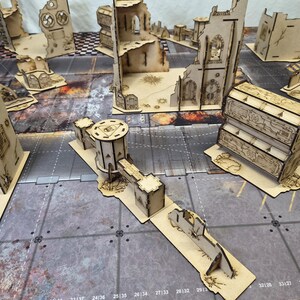 May include: A collection of laser-cut wooden miniature terrain pieces for tabletop gaming, including ruined building walls, a multi-level structure, a cylindrical tower with a bridge, and barricades, all featuring intricate engraved details.