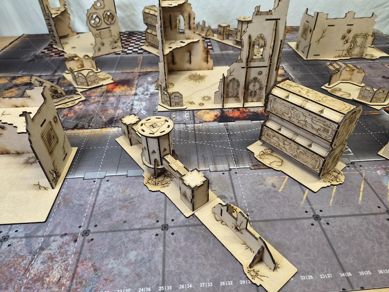 May include: A collection of laser-cut wooden miniature terrain pieces for tabletop gaming, including ruined building walls, a multi-level structure, a cylindrical tower with a bridge, and barricades, all featuring intricate engraved details.