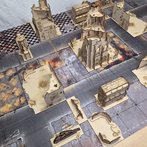 May include: A collection of laser-cut wooden miniature terrain pieces for tabletop gaming, including ruined building structures, barricades, and rectangular supply crates, arranged on a printed game mat with a city street grid pattern.